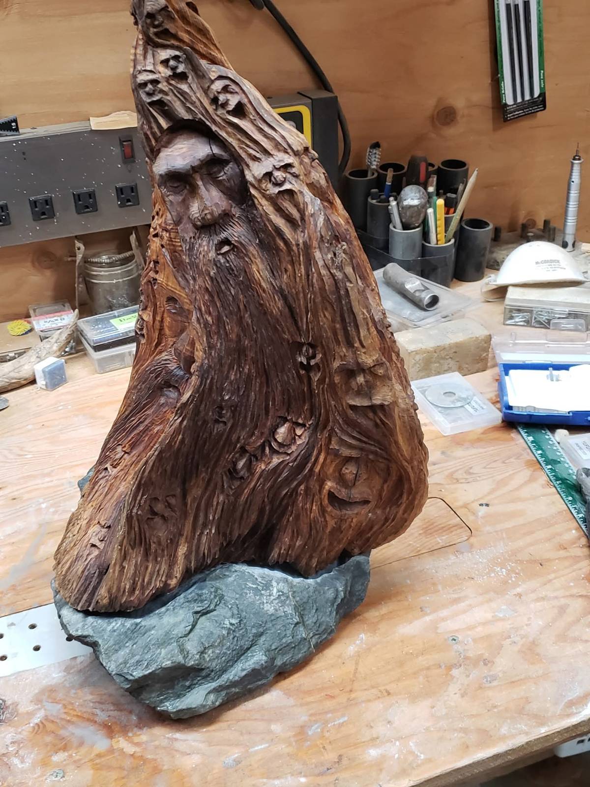Wood spirit carving with a bearded face and dozens of smaller faces, on its stone base